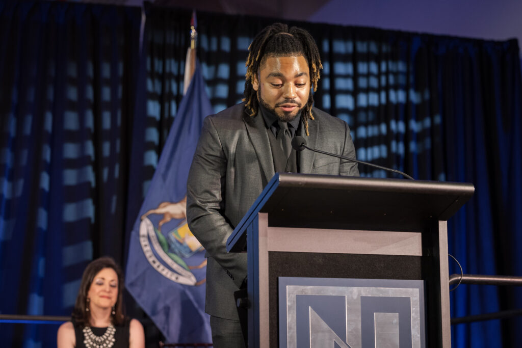 A person in a suit speaks at a podium with a state flag behind them. Another person sits in the background.