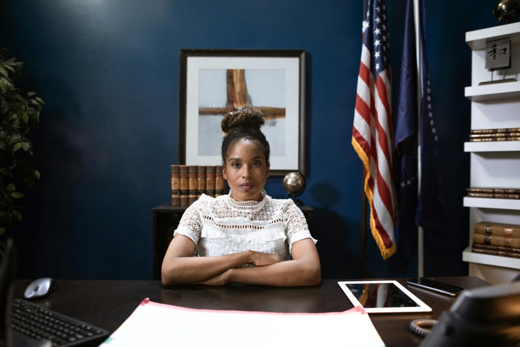 A woman sits at a desk with folded arms, exuding an air of leadership in an office adorned with flags and bookshelves, embodying her dedication to public safety and service.