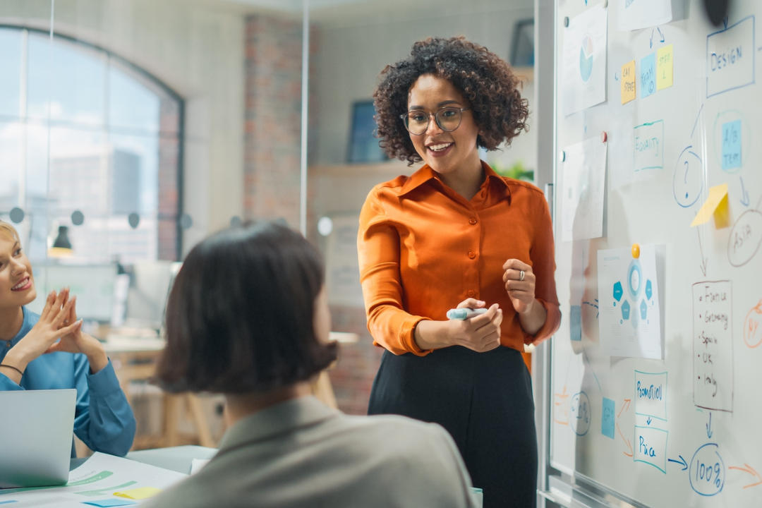 A woman demonstrates leadership as she presents at a whiteboard with charts and sticky notes to colleagues in a modern office. Two people listen attentively, one clapping, while another takes notes.