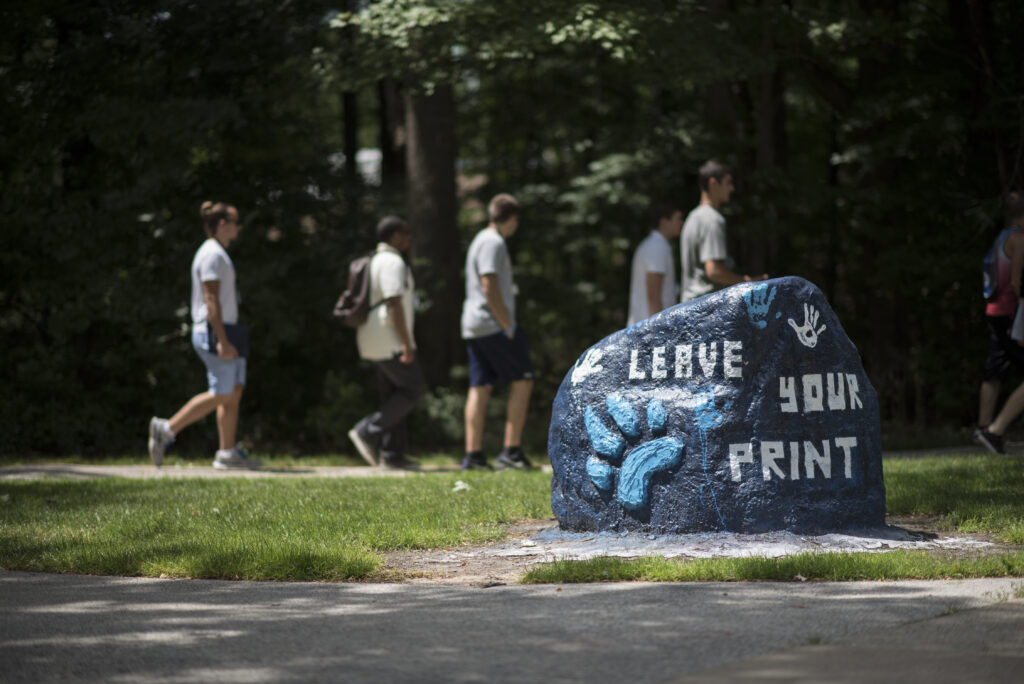 A group of people walk on a path behind a large rock painted with blue paw prints and the words "Leave Your Print.