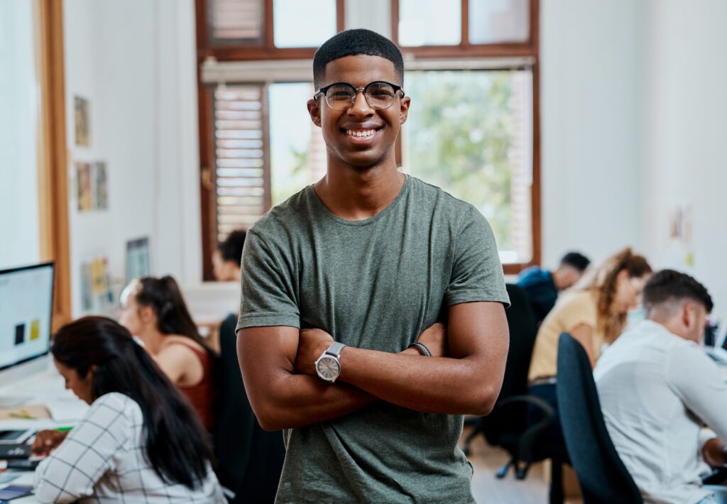 A young man wearing glasses and a green t-shirt stands smiling with arms crossed in a busy, modern office environment.