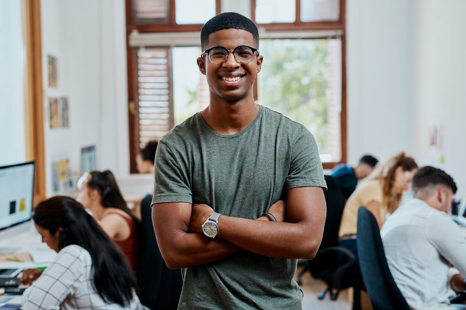 A young man wearing glasses and a green t-shirt stands smiling with arms crossed in a busy, modern office environment.