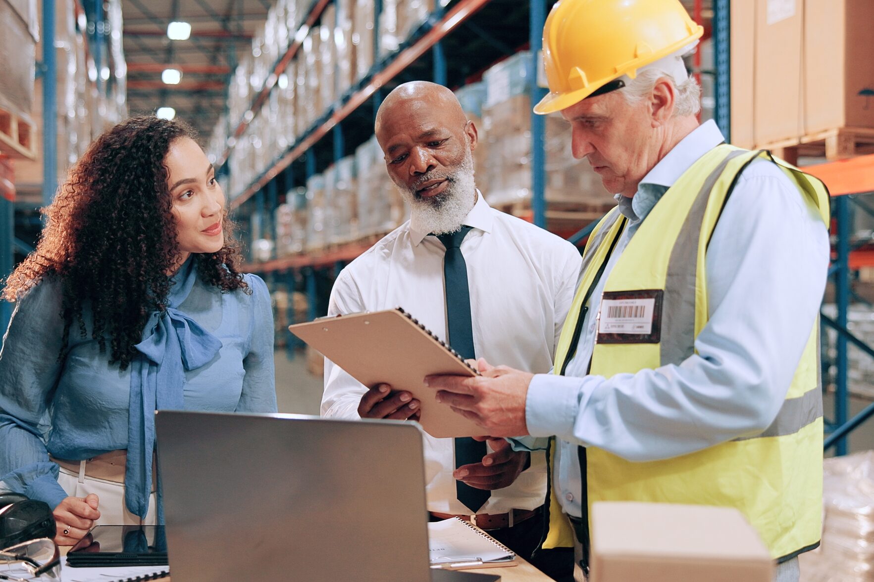 Three people stand in a warehouse discussing documents; one wears a hard hat and safety vest, while the others in business attire review a clipboard, focusing on Lean Operations & Supply Chain Management strategies.